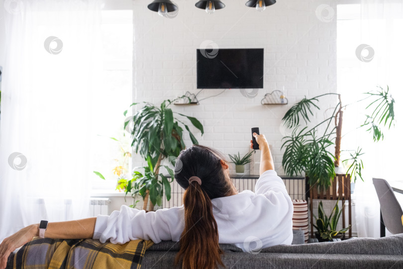 Скачать Женщина дома в белом интерьере сидит на диване и смотрит телевизор, макет черного экрана. Домашний отдых, досуг фотосток Ozero