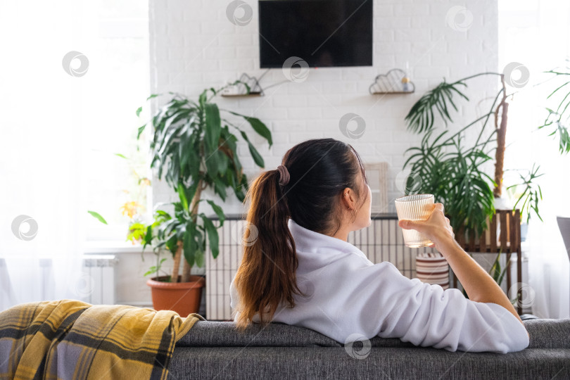 Скачать Женщина дома пьет воду из стакана. в белом интерьере сидит на диване и смотрит телевизор, макет черного экрана. Домашний отдых, досуг фотосток Ozero