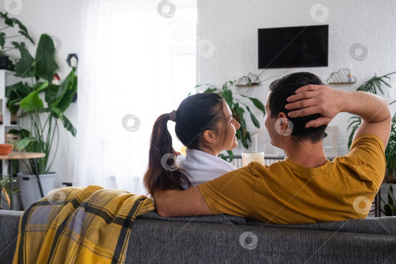 Скачать Мужчина и женщина дома в белом интерьере сидят на диване и смотрят телевизор, макет черного экрана. Домашний отдых, досуг, любовь, семья, свидание фотосток Ozero