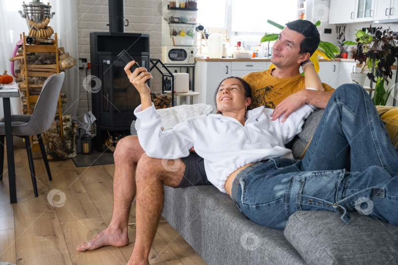 Скачать Мужчина и женщина дома в белом интерьере сидят на диване и смотрят телевизор. Домашний отдых, досуг, любовь, семья, свидание фотосток Ozero