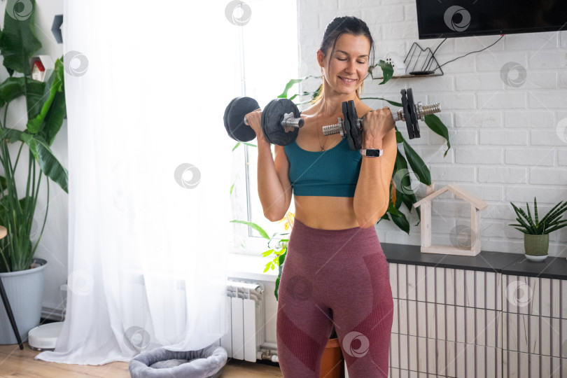 Скачать Женщина в спортивной форме занимается фитнесом и силовыми тренировками со штангой и гантелями у себя дома в интерьере фотосток Ozero