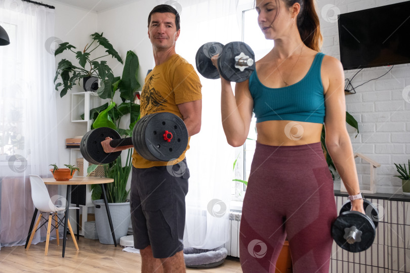 Скачать Женщина и мужчина в спортивной форме занимаются фитнесом и силовыми тренировками со штангой и гантелями дома в интерьере и кат фотосток Ozero