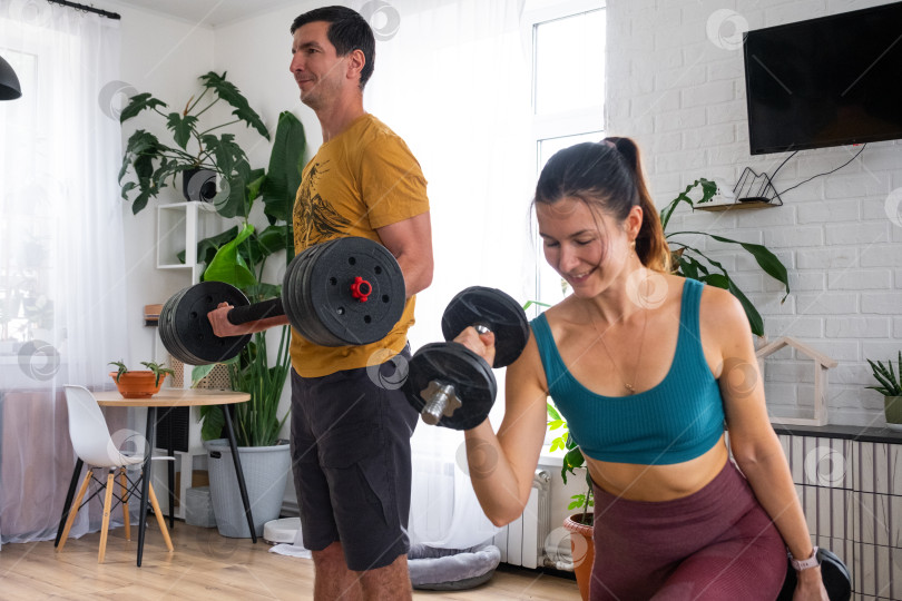 Скачать Женщина и мужчина в спортивной форме занимаются фитнесом и силовыми тренировками со штангой и гантелями у себя дома в интерьере и кат фотосток Ozero