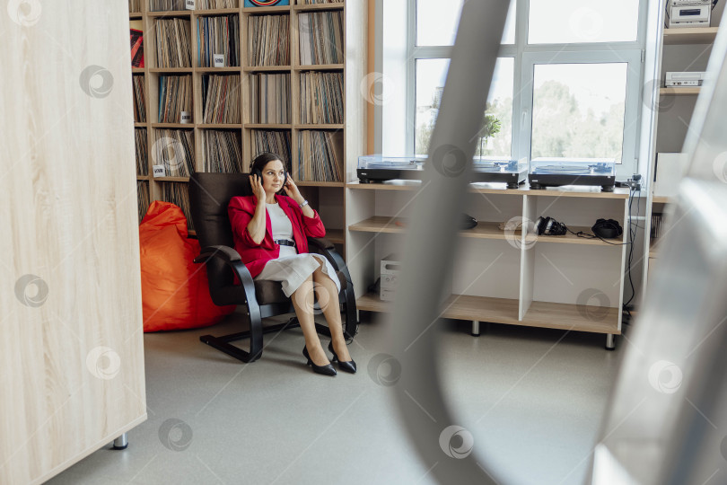 Скачать Молодая азиатка в красном блейзере сидит в современном коворкинге. Она разговаривает по телефону в окружении книжных полок и непринужденной атмосферы. фотосток Ozero