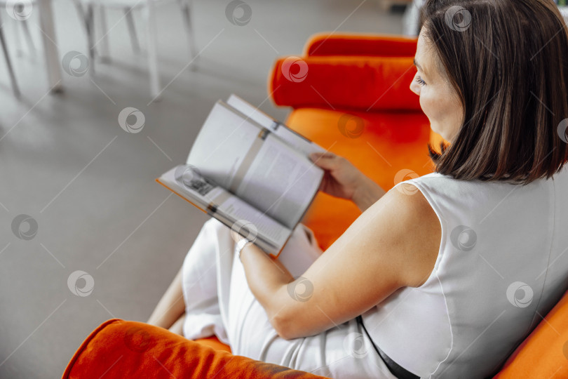 Скачать молодая испаноязычная женщина с короткими каштановыми волосами сидит на оранжевом диване в библиотеке и читает книгу. На заднем плане видны книжные полки, заполненные книгами. фотосток Ozero