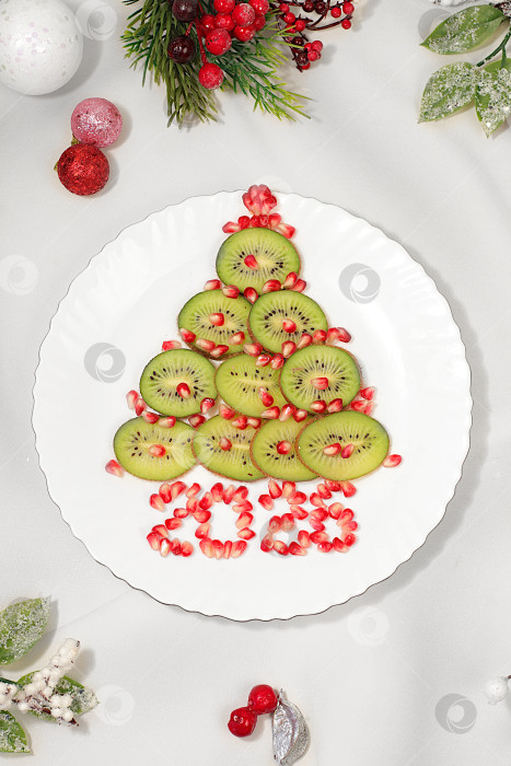 Скачать Зимний фон для поздравительной открытки. Съедобная елка из киви и граната на праздничном столе, идея для оформления новогодних блюд для детей. фотосток Ozero