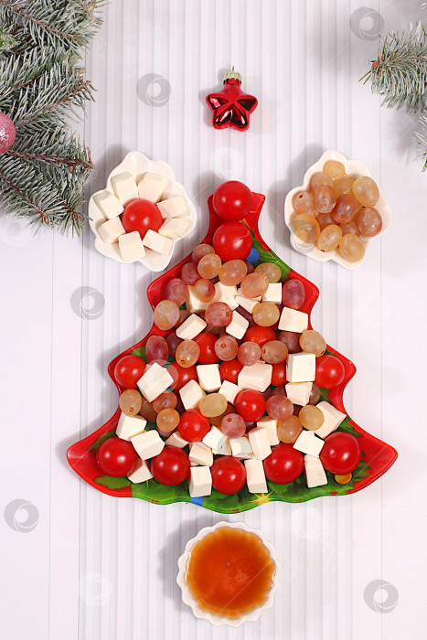 Скачать Праздничный новогодний салат в форме елочки с сыром фета, помидорами, виноградом, оливками и медом, украшенный еловыми ветками и другими украшениями. Идея оформления блюда. Рождественские и новогодние блюда, шаблон меню и баннер ресторана. фотосток Ozero