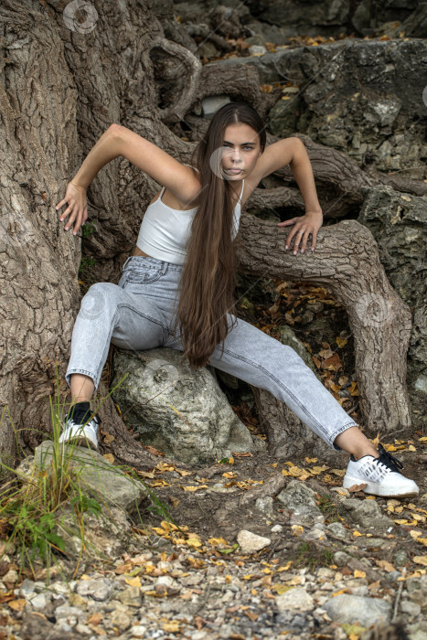 Скачать Молодая красивая женщина в серых джинсах позирует на берегу у корней большого дерева фотосток Ozero