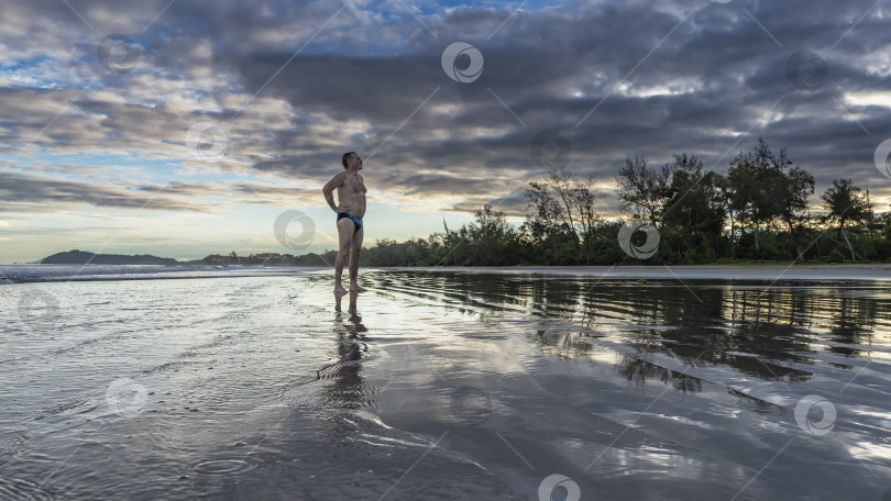 Скачать Мужчина в плавках стоит на песчаном пляже на рассвете. фотосток Ozero