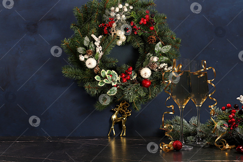 Скачать Шампанское, праздничные украшения и еловый венок со статуэткой оленя на элегантном фоне - концепция кануна Рождества. Новогодняя открытка, баннер для демонстрации, Поздравление с Новым годом фотосток Ozero