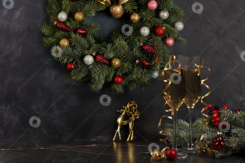 Скачать Шампанское, праздничные украшения и еловый венок со статуэткой оленя на элегантном фоне - концепция кануна Рождества. Новогодняя открытка, баннер для демонстрации, Поздравление с Новым годом фотосток Ozero