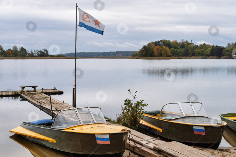 Скачать Золотая осень на Озере Селигер. Природный пейзаж с металлическими лодками казанками и флагом Военно-морского флота на флагштоке у деревянного причала. фотосток Ozero