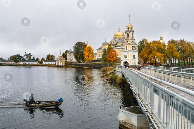 Скачать Величественный вид на Нило-Столбенский монастырь (Нилова пустынь) с моста. Одинокий рыбак на моторной лодке проплывает по озеру Селигер. Осенний пейзаж и спокойствие. фотосток Ozero