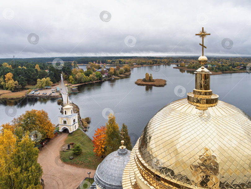 Скачать Величественный панорамный вид с колокольни Нило-Столбенского монастыря на озеро Селигер. Золотой купол на переднем плане, мост-насыпь, маленький островок, изрезанный берег и живописные деревенские дома, окруженные яркими осенними деревьями. фотосток Ozero