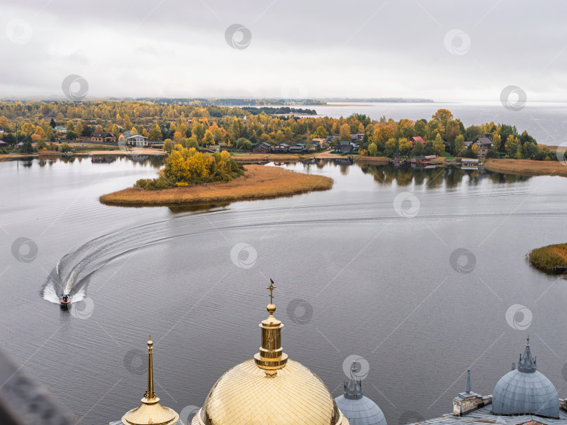 Скачать Величественный панорамный вид с колокольни Нило-Столбенского монастыря на озеро Селигер. Моторная лодка и заметный след на воде в безмятежном заливе. Островок, деревенские дома и живописные осенние деревья. фотосток Ozero