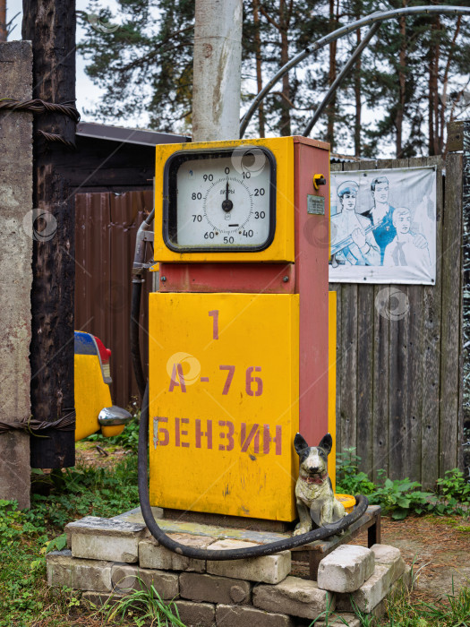 Скачать Ретро бензоколонка А-76 советской эпохи на фоне соснового леса и старого плаката. Крупный вертикальный план исторического объекта. На заднем плане виден старый советский плакат с рабочими и военными. фотосток Ozero