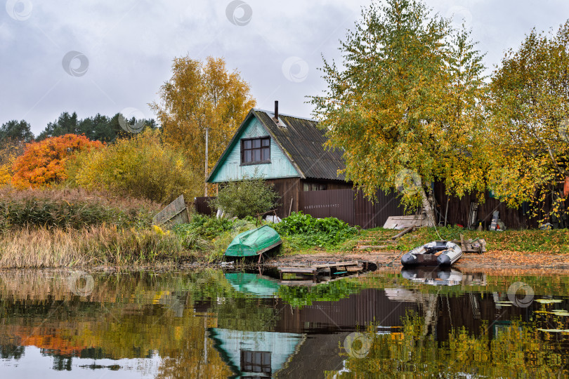 Скачать Пезажный панорамный вид на старый деревянный дом и лодочный причал на берегу озера Селигер. Осенний пейзаж с яркими деревьями и отражением в спокойной воде. фотосток Ozero