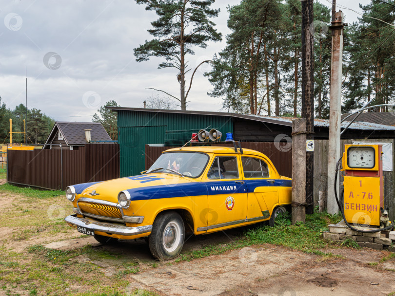 Скачать Ретро милицейский автомобиль Волга (ГАЗ-21) желто-синего цвета. Исторический советский автомобиль на фоне соснового леса и старой бензоколонки. фотосток Ozero