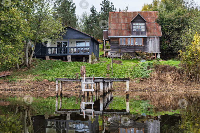 Скачать Панорамный вид на контрастные загородные дома на берегу озера Селигер. Деревянный пирс с лестницей и отражением в спокойной воде. фотосток Ozero