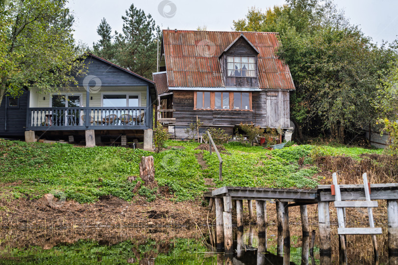 Скачать Панорамный вид на контрастные загородные дома на берегу озера Селигер. Старый деревянный дом со ржавой крышей рядом с современным коттеджем. фотосток Ozero