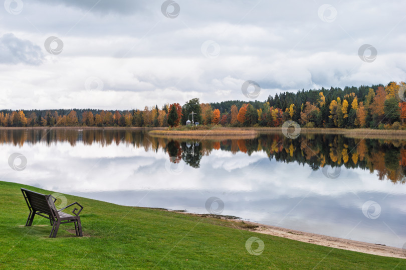 Скачать Панорама озера Селигер с осенним лесом и крестом. Скамейка на зеленом берегу на фоне живописного осеннего озера. Вдали, на узком островке, виден православный крест. Дальний берег покрыт смешанным лесом с яркой желтой, оранжевой и зеленой листвой. Водная гладь зеркально отражает лес и облачное небо фотосток Ozero