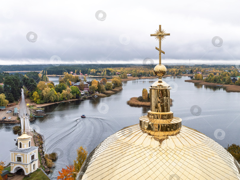 Скачать Величественный панорамный вид с колокольни Нило-Столбенского монастыря на озеро Селигер. Золотой православный крест, Светлицкая башня, мост-насыпь и моторная лодка со следом на воде на фоне живописных осенних деревьев. фотосток Ozero