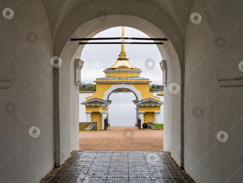 Скачать Величественный вид из арочного проема ворот на Архиерейскую пристань Нило-Столбенского монастыря. Желтое здание часовни с золотым шпилем и безмятежная гладь озера Селигер. фотосток Ozero