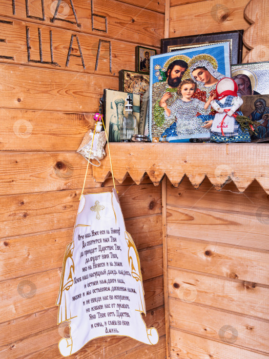 Скачать Крупный план икон и молитвы "Отче наш" в деревянной часовне на озере Селигер. На деревянной полке расположены разнообразные иконы, включая образ Святого Семейства. фотосток Ozero