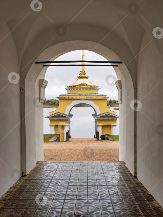 Скачать Крупный вертикальный вид из арочного проема монастырских ворот на Архиерейскую пристань Нило-Столбенского монастыря. Желтое здание часовни с золотым шпилем и безмятежная гладь озера Селигер. фотосток Ozero
