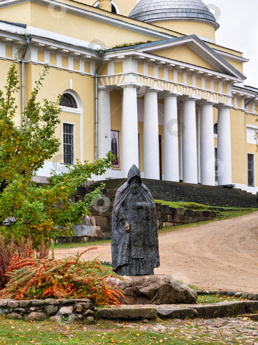 Скачать Крупный план памятника преподобному Нилу Столобенскому на огромном камне-валуне в Ниловой пустыни. Бронзовая скульптура на фоне колоннады Преображенского собора. фотосток Ozero