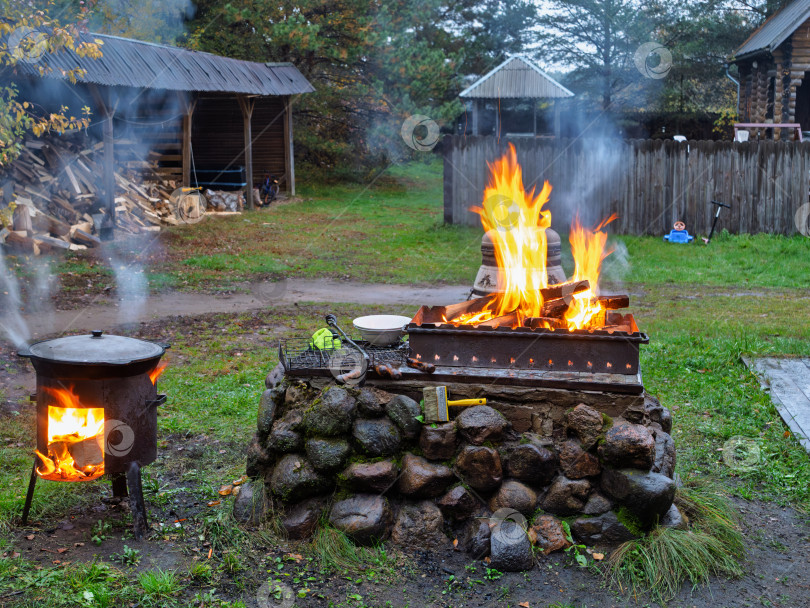 Скачать Мангал и казан с огнем на заднем дворе деревенского дома. Приготовление еды на открытом воздухе. Слева — чугунный казан на отдельной печи, из которого идет пар. В центре — мангал на каменном основании, в котором ярко горит огонь. На заднем плане слева видна дровница с большим запасом сложенных дров, а справа — деревянный забор и постройки. фотосток Ozero