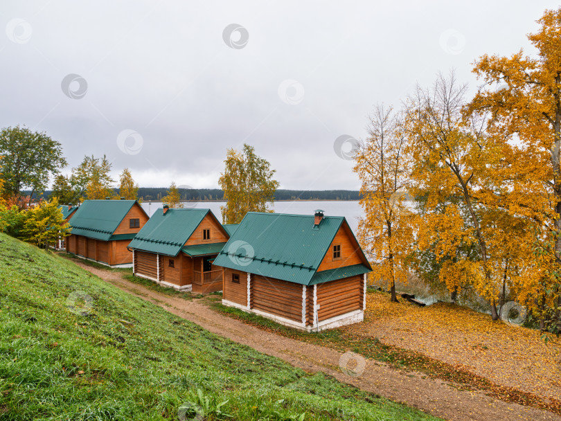 Скачать Деревянные домики для паломников на склоне холма у озера Селигер. Осенние березы с золотистой листвой и зеленые крыши домов создают живописный пейзаж. фотосток Ozero