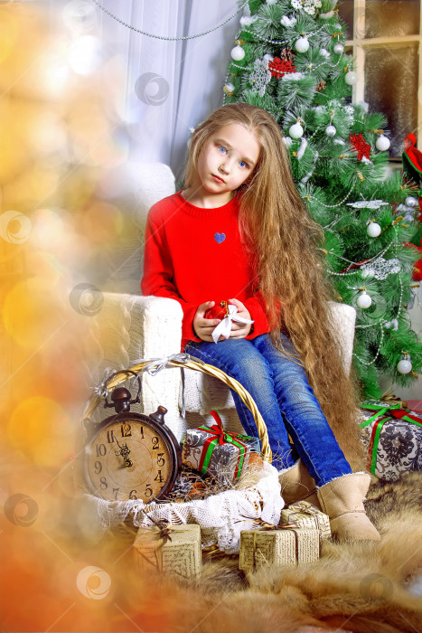 Скачать Девочка- ребенок с распущенными вьющимися волосами и в красной куртке, сидящая на белом диване. На заднем плане рождественская елка, украшенная игрушками. Выборочный фокус фотосток Ozero