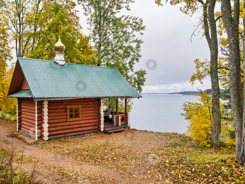 Скачать Уединенная деревянная часовня-скит на высоком берегу озера Селигер в Ниловой пустыни. Зеленая крыша, золотой купол и осенние деревья создают атмосферу покоя. фотосток Ozero
