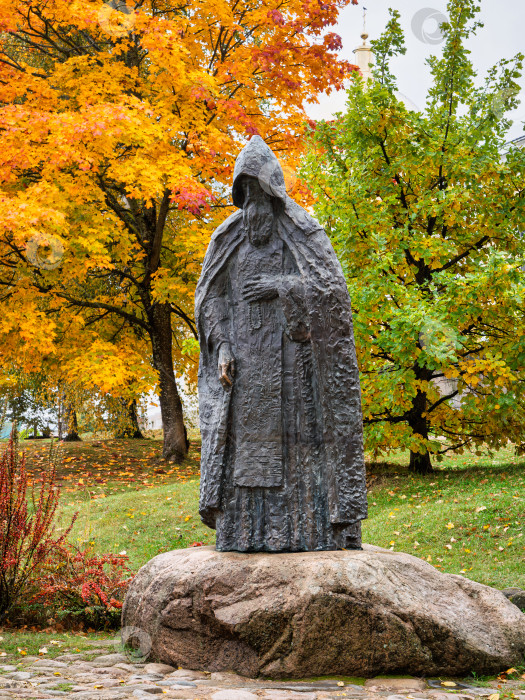 Скачать Портретный вид памятника преподобному Нилу Столобенскому на огромном камне-валуне в Ниловой пустыни. Бронзовая скульптура на фоне яркой золотой осенней листвы. фотосток Ozero