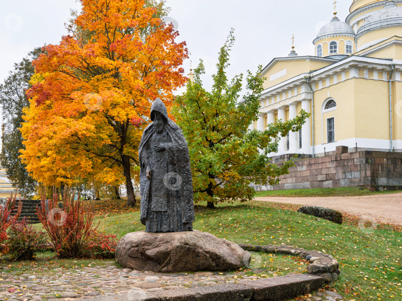 Скачать Портретный вид памятника преподобному Нилу Столобенскому на огромном камне-валуне в Ниловой пустыни. Бронзовая скульптура на фоне яркой золотой осенней листвы и Преображенского собора. фотосток Ozero