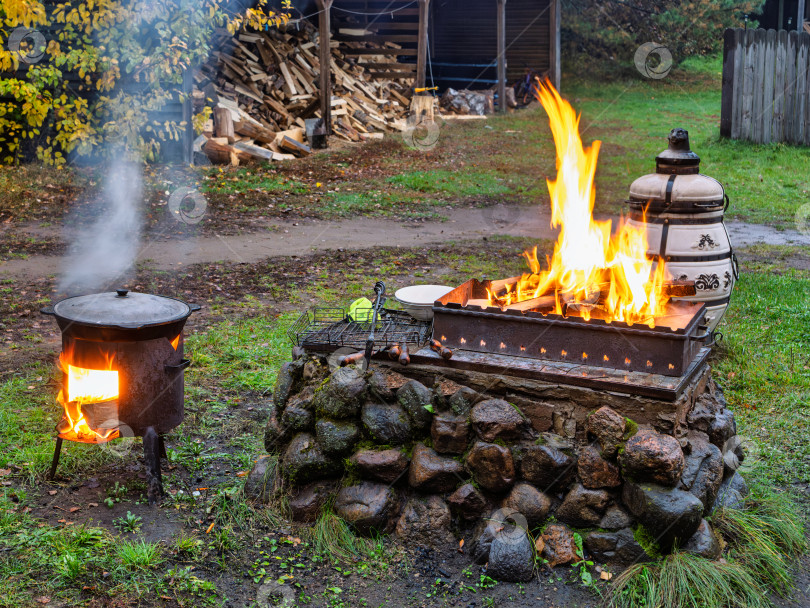 Скачать Зона для барбекю и тандыр с огнем. Приготовление еды на открытом воздухе на озере Селигер. В центре — мангал на основании из камней, в котором ярко горит огонь. Слева — чугунный казан на отдельной печи, из которого идет пар. На заднем плане — дровница с аккуратно сложенными дровами. фотосток Ozero