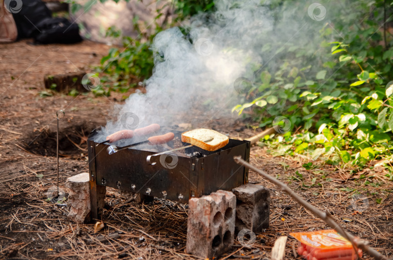 Скачать Сосиски и хлеб жарятся на мангале в лесу, много дыма фотосток Ozero