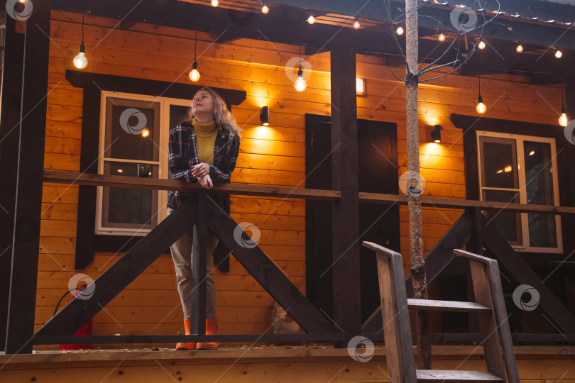 Скачать Женщина в просторном уютном свитере и сапогах наслаждается золотым часом на веранде в деревенском стиле в окружении природы. Тихий и безмятежный момент на свежем воздухе, охватывающий простоту фотосток Ozero