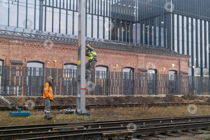 Скачать Рабочие устанавливают систему оповещения на столбе на железной дороге на фоне промышленных зданий. фотосток Ozero