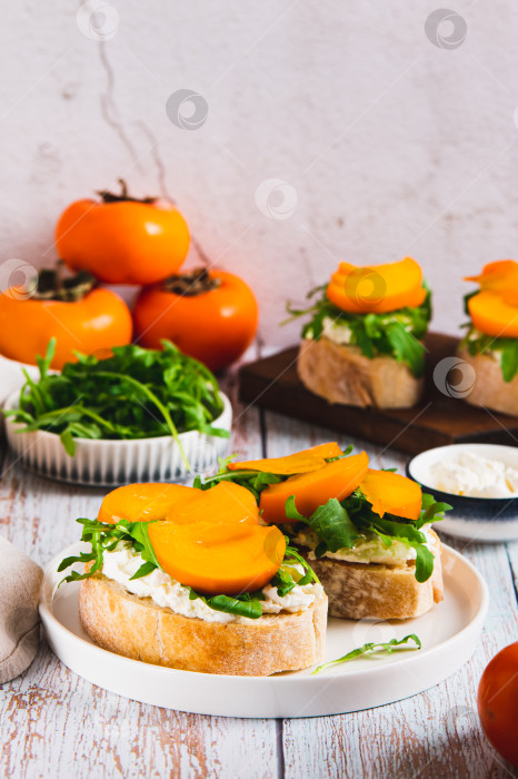 Скачать Сладкие бутерброды с рикоттой, рукколой и хурмой на тарелке на столе в вертикальном положении фотосток Ozero