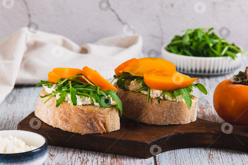 Скачать Хурма, руккола и сливочный сыр в чиабатте на деревянной доске на столе фотосток Ozero