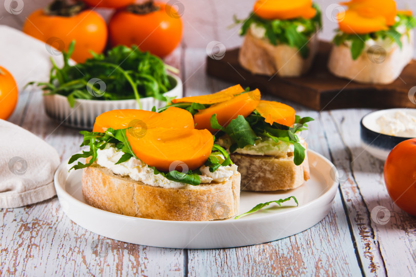 Скачать Сладкие бутерброды с рикоттой, рукколой и хурмой на тарелке на столе фотосток Ozero
