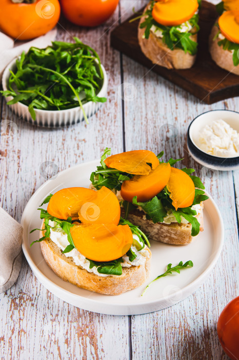 Скачать Чиабатта с хурмой, зеленью и сливочным сыром на тарелке на столе, вид сверху фотосток Ozero