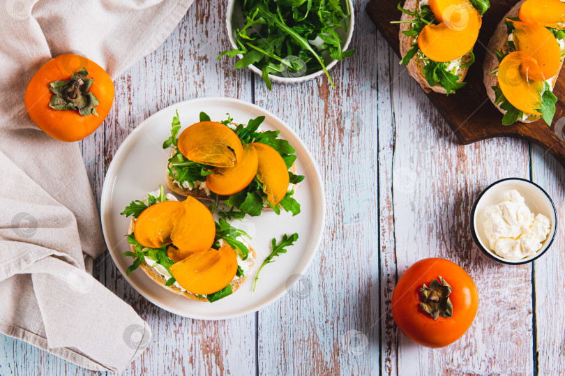 Скачать Сладкие бутерброды с рикоттой, рукколой и хурмой на тарелке на столе вид сверху фотосток Ozero