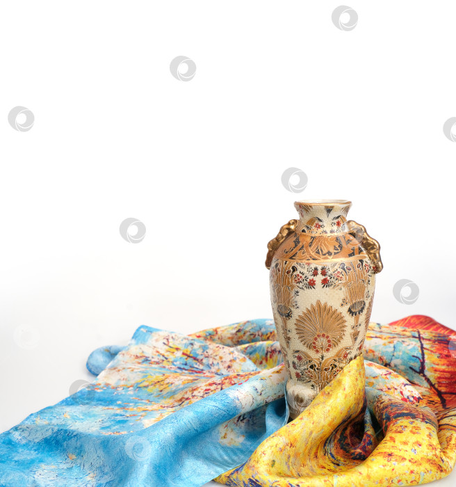 Скачать Декоративная керамическая ваза с замысловатыми золотыми и коричневыми цветочными узорами стоит на ложе из разноцветных шелковых шарфов фотосток Ozero