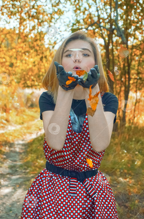 Скачать Девушка в ретро-платье в горошек стоит и дует на ладони, из ее рук вылетают сухие листья. Сельская местность, тропинка в лесу. Летнее солнечное утро фотосток Ozero