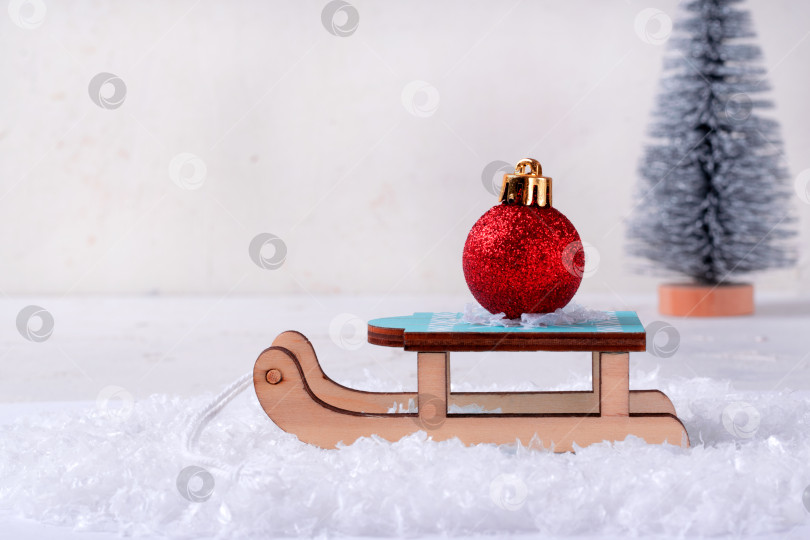 Скачать Красный елочный шар на игрушечных деревянных санках в искусственном снегу фотосток Ozero