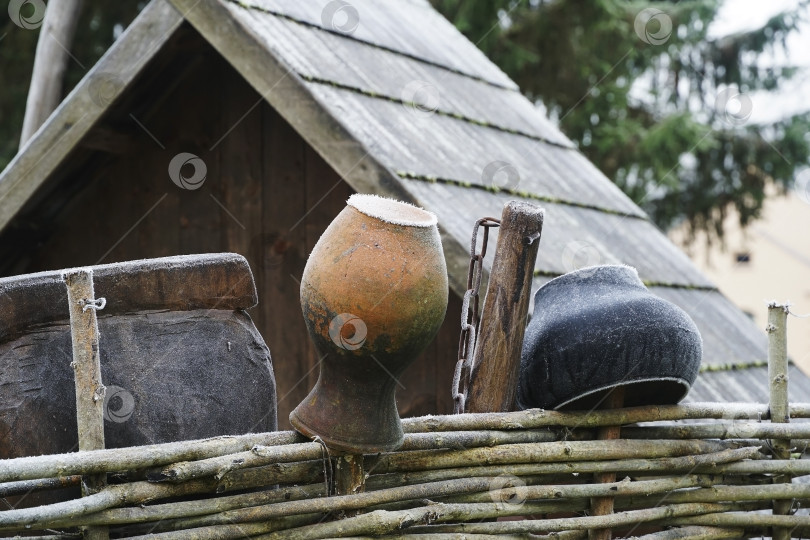 Скачать Сельский пейзаж. Крынка и чугунный горшок на сельском плетне морозным утром. На заднем плане деревянное корыто и колодец. фотосток Ozero