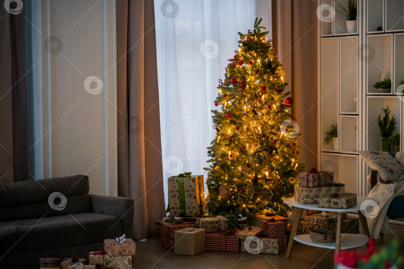 Скачать Украшенная рождественская елка со светящимися огнями и подарочной коробкой под ней в гостиной. фотосток Ozero